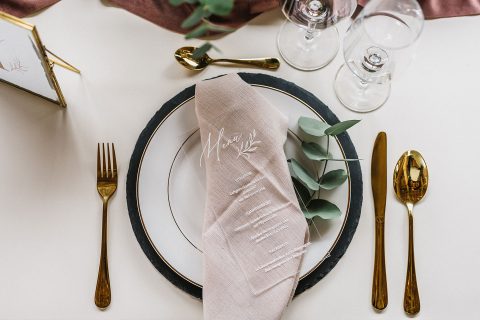 Hochzeitswahn - Elegantes Tischgedeck mit einem schwarzen Teller mit Goldrand, einer beigen Leinenserviette mit Kursivschrift, goldenem Besteck und Kristallgläsern auf einer kastanienbraunen Tischdecke mit grünen Akzenten. Magical-Homes