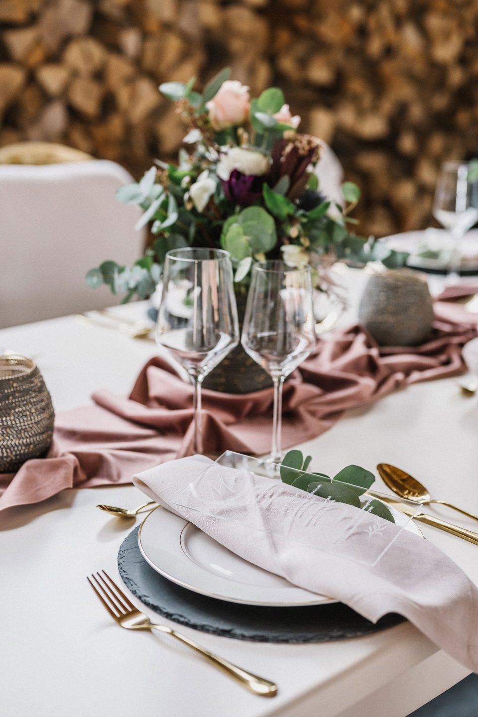 Hochzeitswahn - Elegantes Esstischgedeck mit goldenem Besteck, einem Stapel Teller, Weingläsern und einem Blumengesteck auf einem Tisch mit blassrosa Tischdecke vor einer strukturierten Holzwand. Magical-Homes