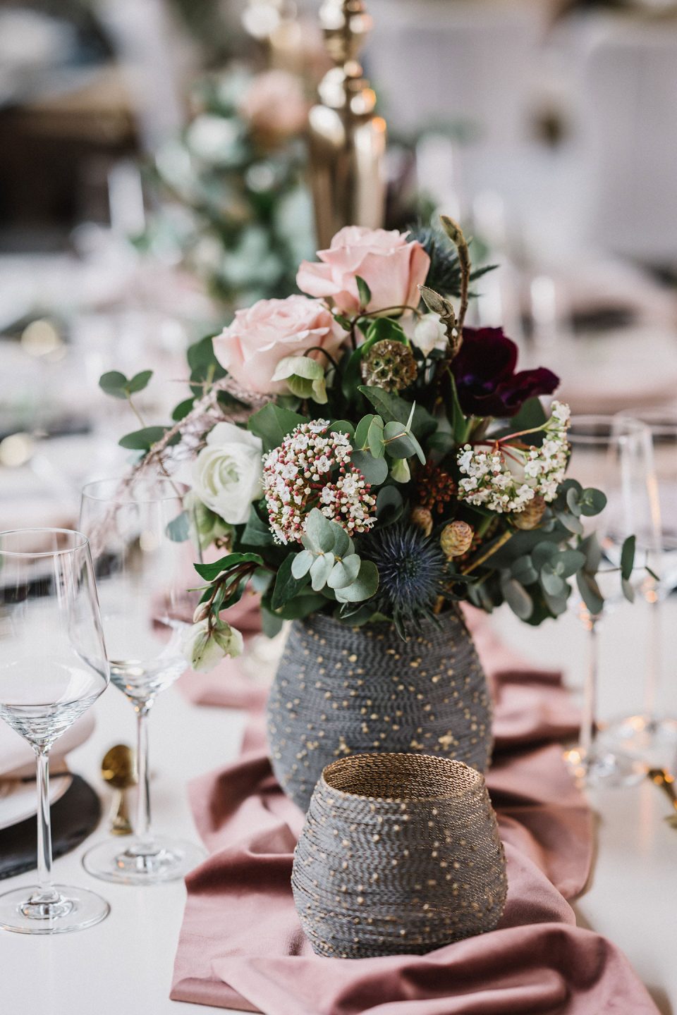 Hochzeitswahn - Elegante Esstischdekoration mit einem Blumenarrangement aus rosa Rosen und grünem Laub in einer strukturierten grauen Vase, ergänzt durch feines Glasgeschirr und zartrosa Tischwäsche. Magical-Homes