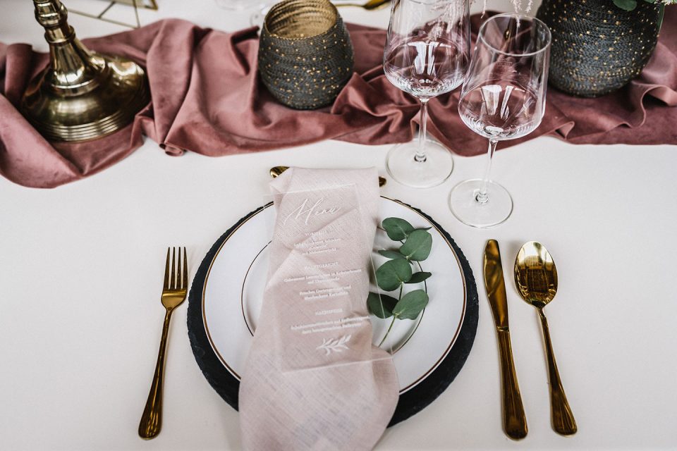 Hochzeitswahn - Elegantes Esstischgedeck mit einem schwarz-weißen Teller mit Goldrand, dazu goldenes Besteck, Weingläser und Dekorationsgegenstände, akzentuiert mit einer rosa Serviette und grünen Blättern. Magical-Homes
