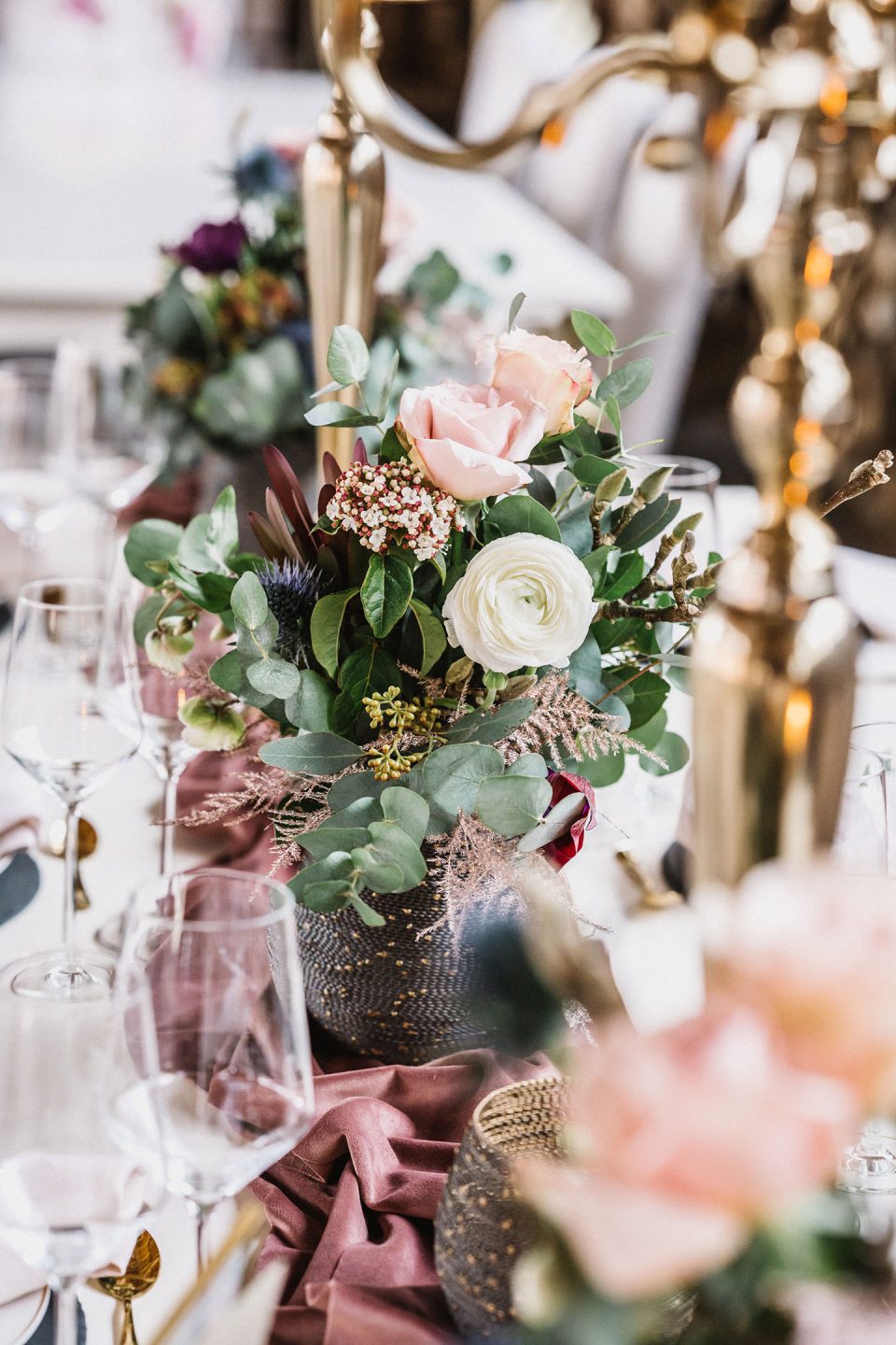 Hochzeitswahn - Elegant arrangierter Esstisch mit floralen Tafelaufsätzen aus rosa und weißen Blumen, grünem Laub in einer dunklen Vase, umgeben von Weingläsern und goldenem Besteck, akzentuiert durch einen dunkelroten Tischläufer. Magical-Homes