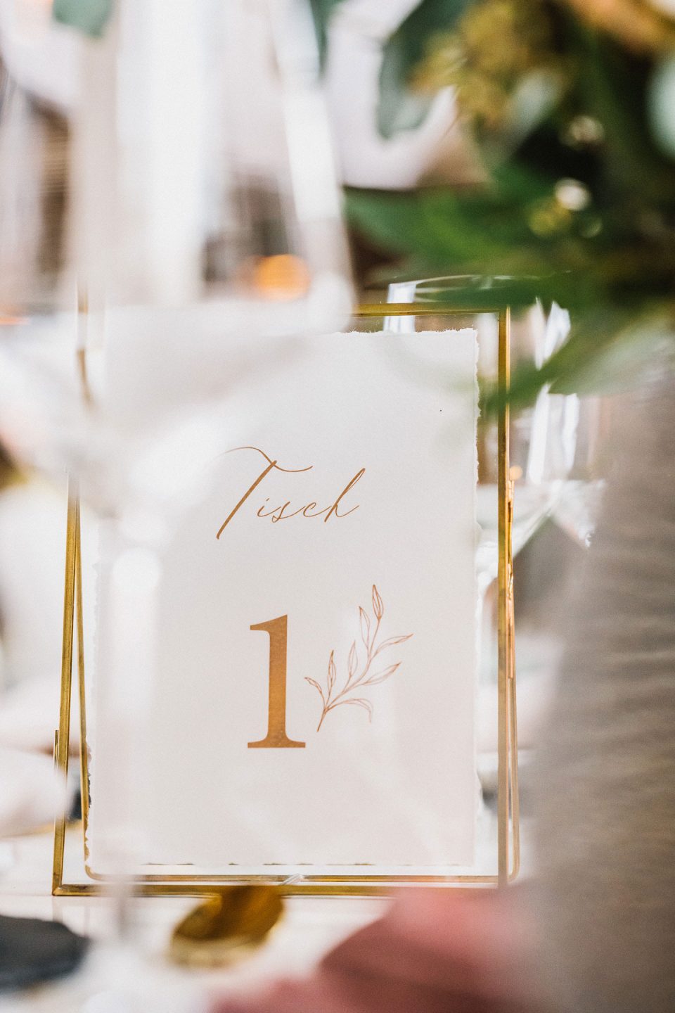 Hochzeitswahn - Elegantes Tischnummernschild „Tisch 1“ mit Schreibschrift und einfacher botanischer Illustration auf transparentem Papier, präsentiert in einem Metallhalter mit weichem, unscharfem Hintergrund. Magical-Homes