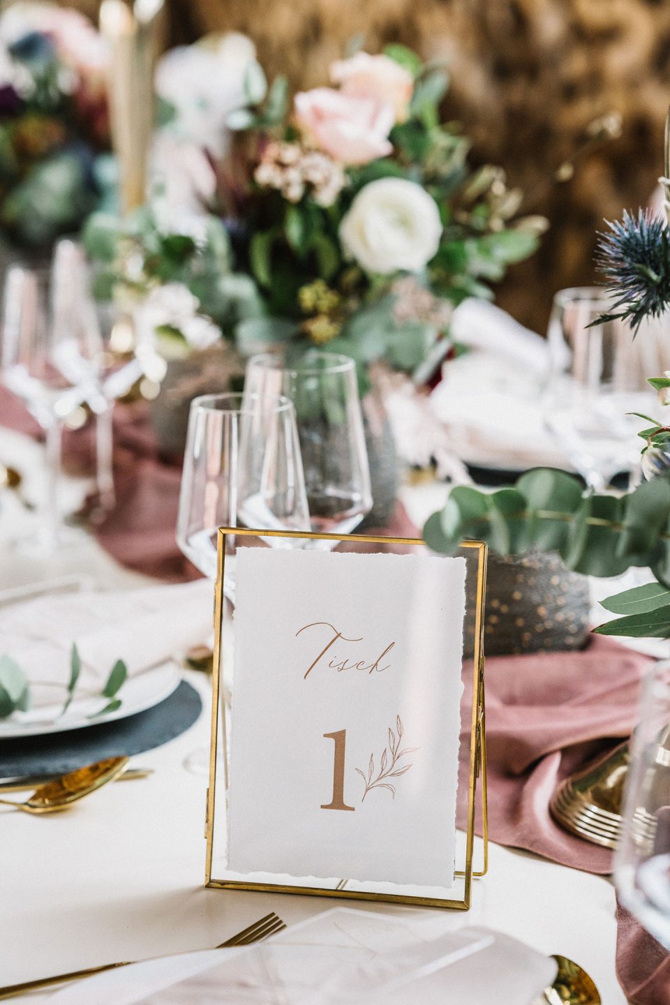 Hochzeitswahn - Elegante Tischdekoration für eine Hochzeit mit einer Tischnummernkarte mit Goldrand und Blumenmuster, umgeben von Kristallgläsern, pastellfarbenen Blumenarrangements und Grünpflanzen auf einer strukturierten Tischdecke. Magical-Homes