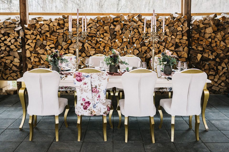 Hochzeitswahn - Ein elegant gedeckter Esstisch mit weißen Stühlen und Blumenmuster, geschmückt mit Kerzen und Blumengestecken, positioniert vor einem Holzstapel in rustikalem Ambiente. Magical-Homes