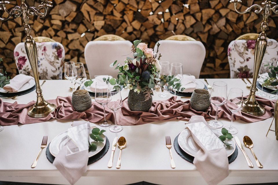 Hochzeitswahn - Elegant arrangierter Esstisch mit floralem Tafelaufsatz, goldenem Besteck und Weingläsern, ergänzt durch dunkelrosa Tischläufer und verzierte Kerzenhalter, vor einem Hintergrund aus gestapeltem Holz. Magical-Homes
