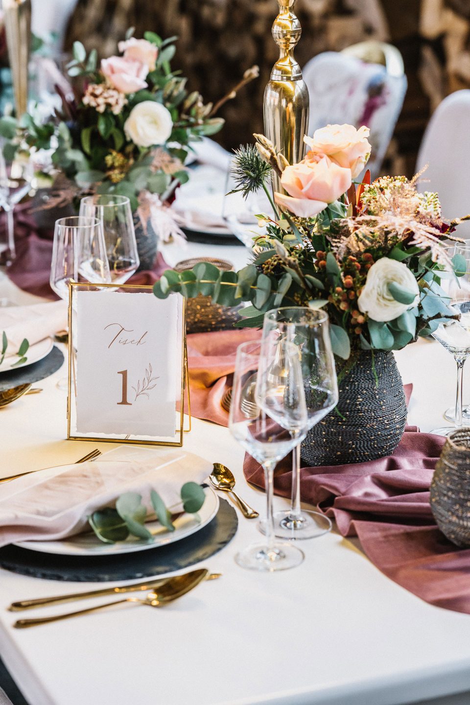 Hochzeitswahn - Elegante Bankett-Tischdekoration mit floralen Tafelaufsätzen, goldenem Besteck, Weingläsern und Speisekarten mit Tischnummer und Blätteremblem. Farbenfrohe Tischdecken ergänzen das anspruchsvolle Dekor. Magical-Homes