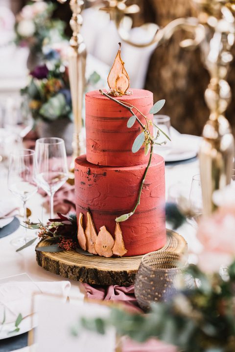 Hochzeitswahn - Eine dreistöckige rote Hochzeitstorte, mit Blattgold und Grün verziert, auf einer Holzplatte in der Mitte, umgeben von elegantem Tischgedeck. Magical-Homes