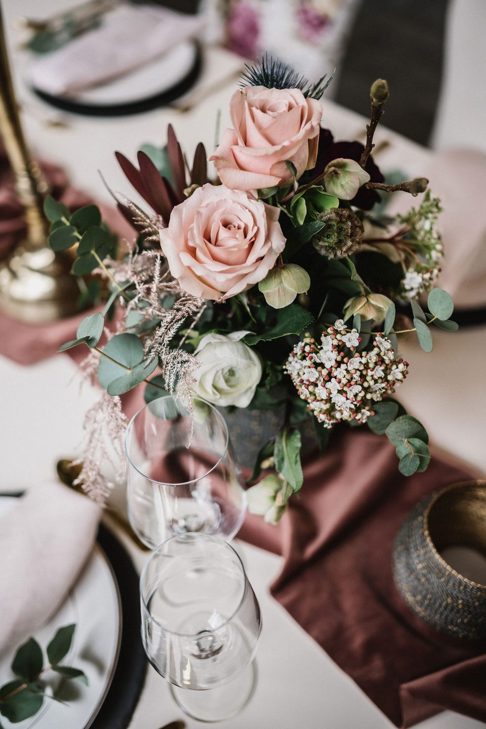 Hochzeitswahn - Eine Nahaufnahme eines elegant gedeckten Tisches mit einem floralen Tafelaufsatz aus rosa Rosen und Grünpflanzen, Weingläsern aus Kristall und feinem Porzellan auf einem mit einer roten Tischdecke geschmückten Tisch. Magical-Homes