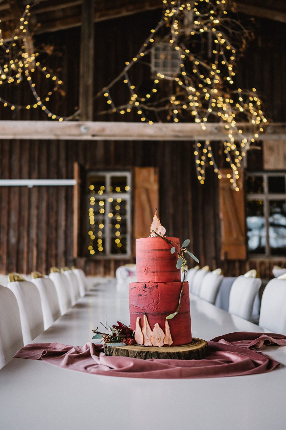 Hochzeitswahn - Eine dreistöckige rote Hochzeitstorte mit goldener Spitze auf einem mit einem zartrosa Tuch bedeckten Holztisch in einer rustikalen Scheune, die mit funkelnden Lichtern geschmückt ist. Magical-Homes