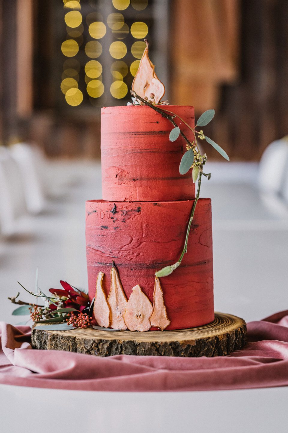 Hochzeitswahn - Eine dreistöckige Hochzeitstorte mit strukturierter roter Glasur, verziert mit goldenen Blättern und kleinen roten Beeren, präsentiert auf einem Holzstumpf und geschmückt mit einem rosa Satinband. Magical-Homes