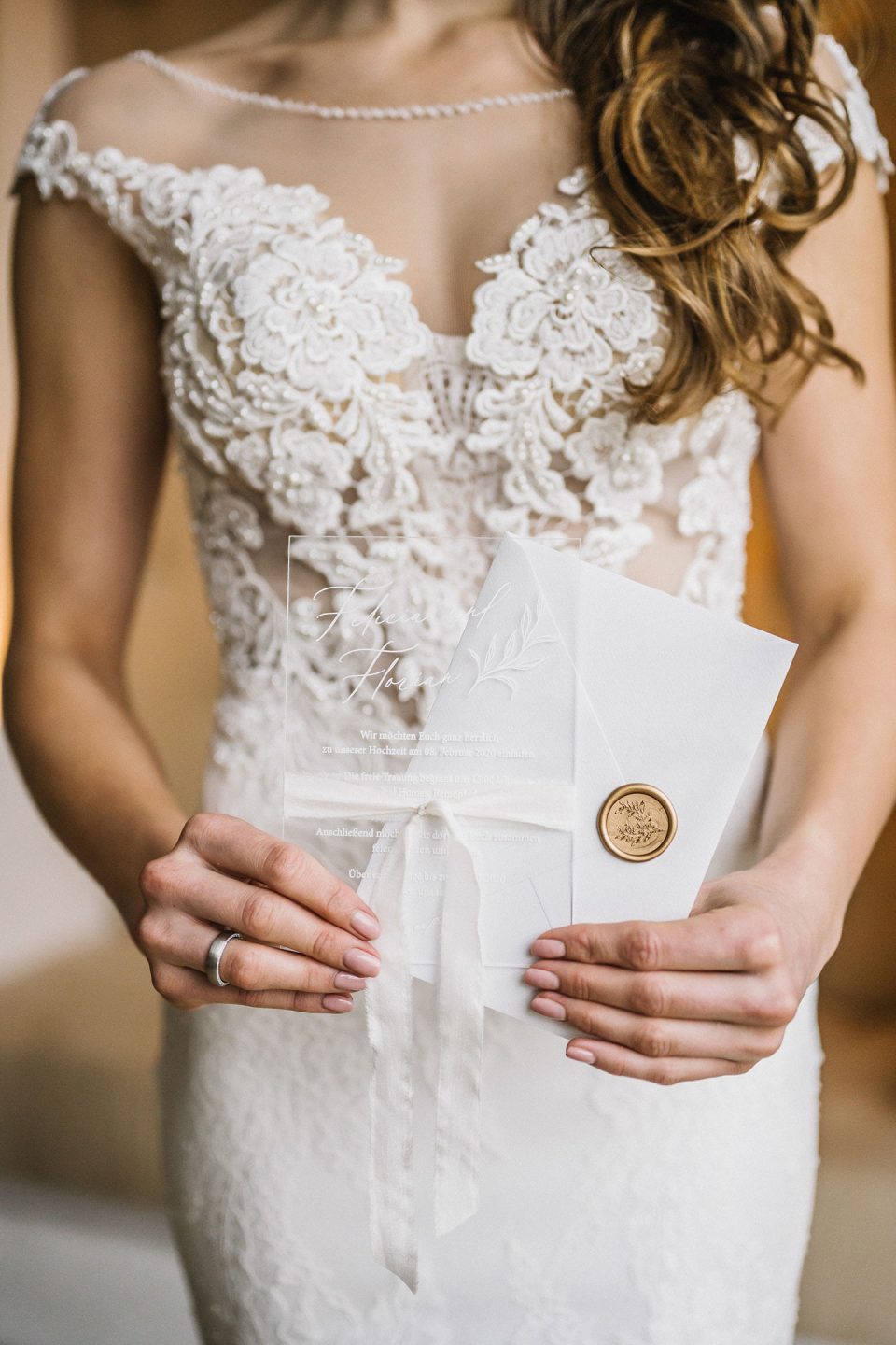 Hochzeitswahn - Eine Braut in einem verzierten Spitzenkleid hält eine Hochzeitseinladung mit einem Wachssiegel und einer weißen Schleife. Nur ihr Oberkörper und ihre Hände sind sichtbar, was die komplizierten Details ihres Kleides und der Einladung betont. Magical-Homes