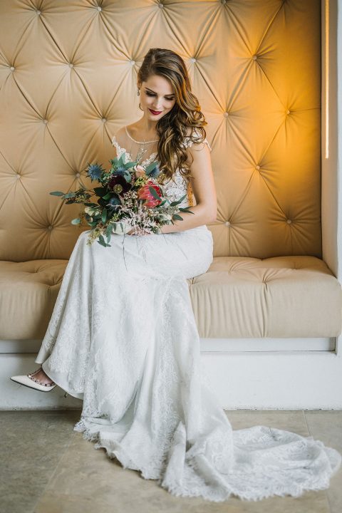 Hochzeitswahn - Eine Braut in einem eleganten weißen Spitzenkleid sitzt auf einer cremefarbenen Bank und hält einen Strauß gemischter Blumen. Im Hintergrund ist eine gemusterte gelbe Wand zu sehen. Magical-Homes