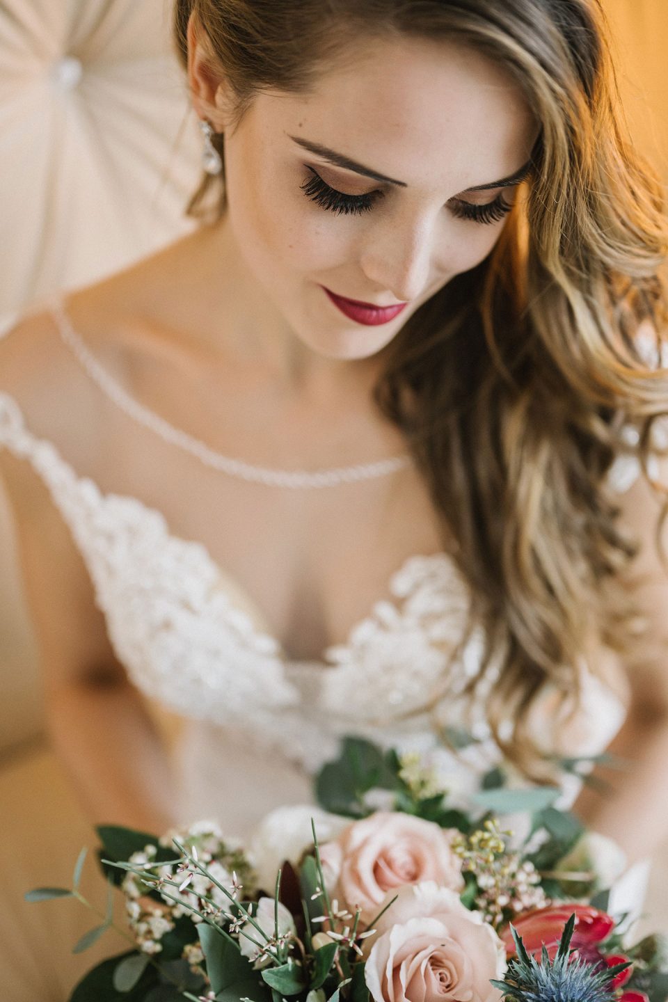 Hochzeitswahn - Eine Braut in einem Spitzenbrautkleid hält einen Rosenstrauß und blickt mit einem sanften Lächeln nach unten. Sie steht unter sanftem Licht, das ihr detailliertes Make-up und ihre elegante Frisur hervorhebt. Magical-Homes