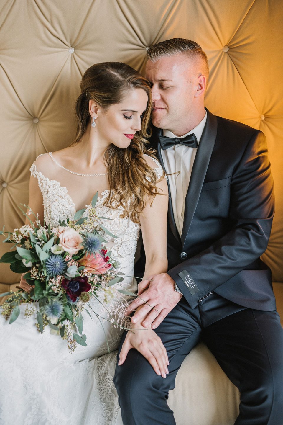 Hochzeitswahn - Eine Braut in einem detailreichen Spitzenkleid und ein Bräutigam im schwarzen Smoking sitzen eng beieinander und teilen einen zärtlichen Moment vor einem reich verzierten, getufteten Hintergrund. Die Braut hält einen Strauß verschiedener Blumen. Magical-Homes