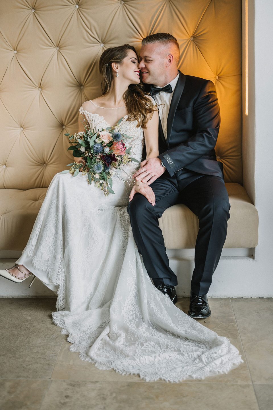 Hochzeitswahn - Eine Braut in einem eleganten Spitzenbrautkleid und ein Bräutigam im schwarzen Anzug sitzen liebevoll zusammen auf einem gepolsterten cremefarbenen Sofa und halten einen großen Blumenstrauß in den Händen. Magical-Homes