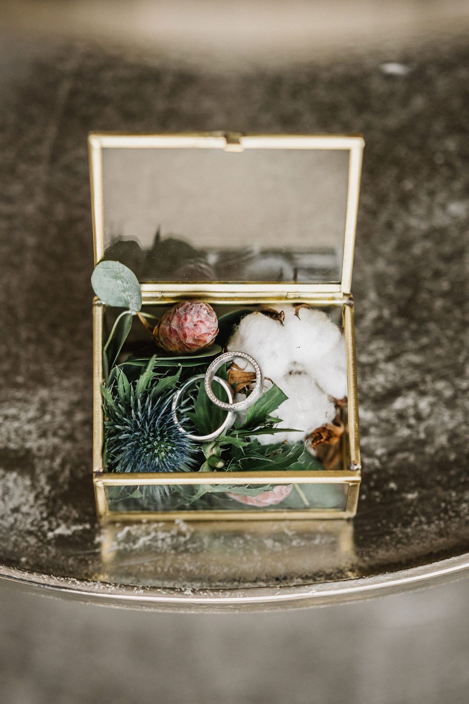 Hochzeitswahn - Eine Glasbox gefüllt mit botanischen Elementen und einem Ehering, platziert auf einer Betonoberfläche. Die Box enthält ein Wattestäbchen, verschiedene Blätter und eine blaue Distel. Magical-Homes