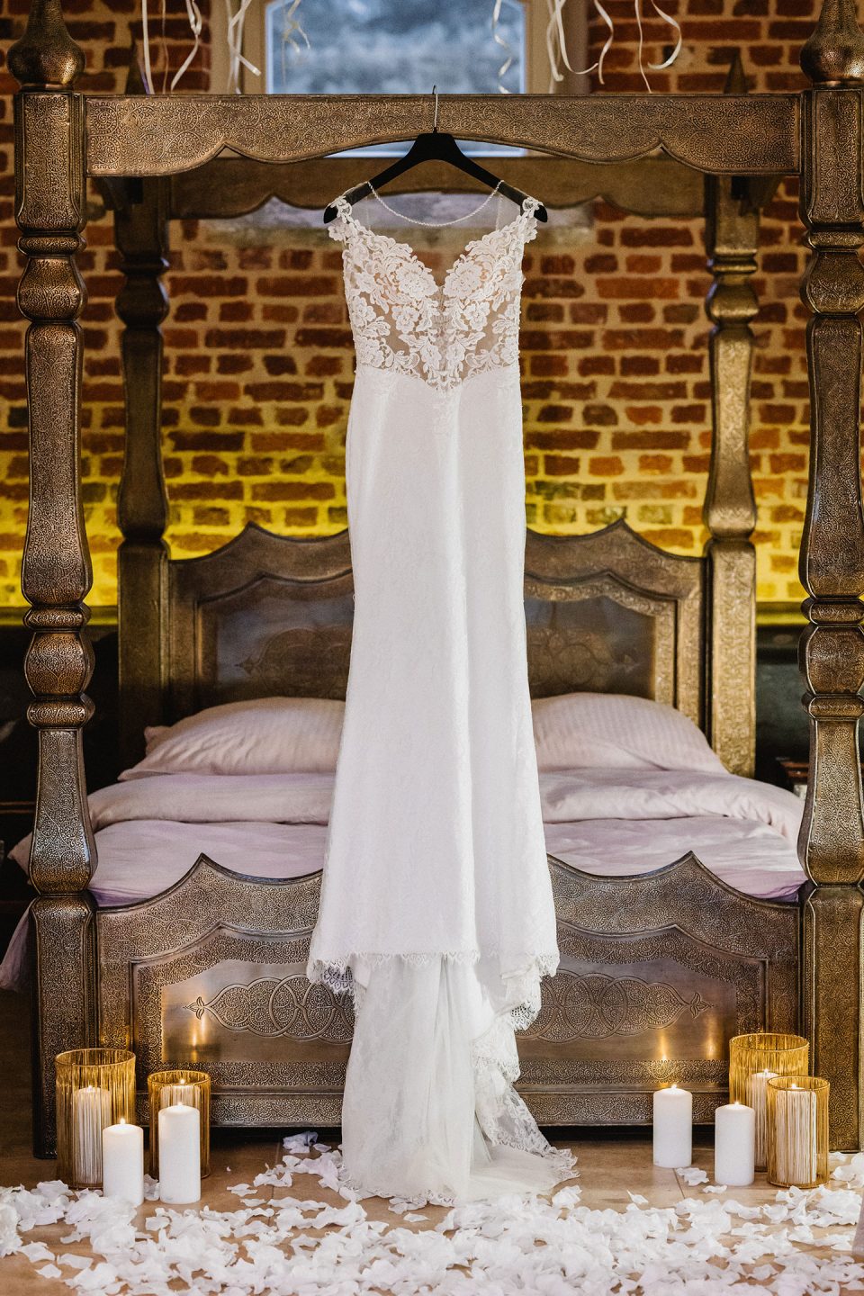 Hochzeitswahn - Ein Spitzenbrautkleid hängt an einem verzierten Bettrahmen aus Holz. Das Bett ist von brennenden Kerzen und verstreuten Rosenblättern umgeben, was eine romantische Atmosphäre schafft. Magical-Homes