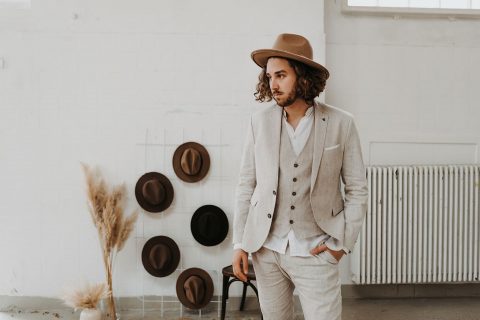Hochzeitswahn - Ein stilvoller Mann in beigem Anzug und braunem Hut posiert nachdenklich neben einer weißen Wand, die mit einer Hutsammlung geschmückt ist, die Hände lässig in den Taschen gesteckt.