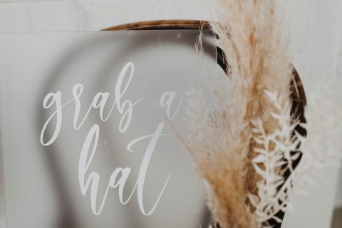 Hochzeitswahn - Ein skurriles kalligrafisches Schild mit der Aufschrift „Schnapp dir einen Hut“ spiegelt sich in einem Spiegel, ergänzt durch die weiche, federleichte Textur von Pampasgras in einer rustikalen Umgebung.