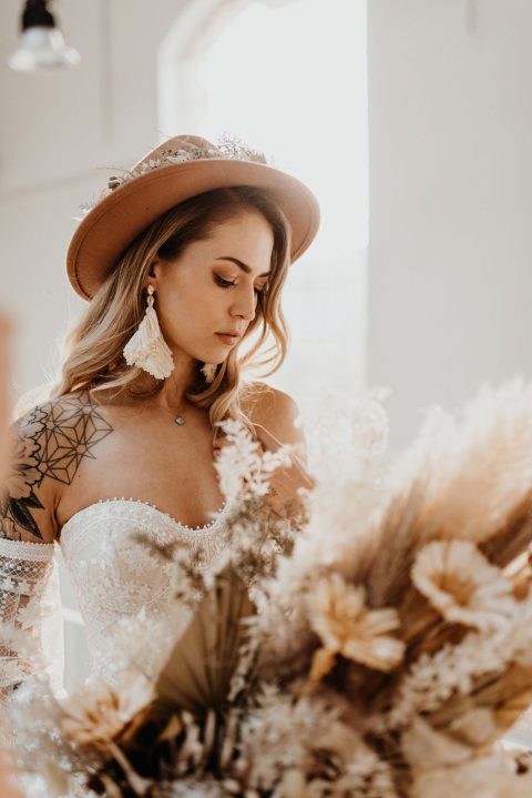 Hochzeitswahn - Eine nachdenkliche Frau in einem Spitzenbrautkleid und einem eleganten Hut blickt nach unten, hält einen Strauß getrockneter Blumen in der Hand und wird von Sonnenlicht sanft beleuchtet.