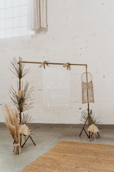 Hochzeitswahn - Eine verträumte Hochzeitskulisse im Bohème-Stil mit einem „Lasst das Abenteuer beginnen“-Schild, einem runden Makramee-Kunstwerk, getrockneten Palmblättern und Pampasgras-Arrangements, auf einem Juteteppich in einem Raum mit weißen Wänden.