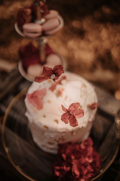 Hochzeitswahn - Ein kleiner, runder Kuchen mit weißem Zuckerguss und rosa getrockneten Blumen, präsentiert auf einem Glaskuchenständer, umgeben von zartrosa Blütenblättern, verströmt herbstliche Verspieltheit. Eine mehrstöckige Portion