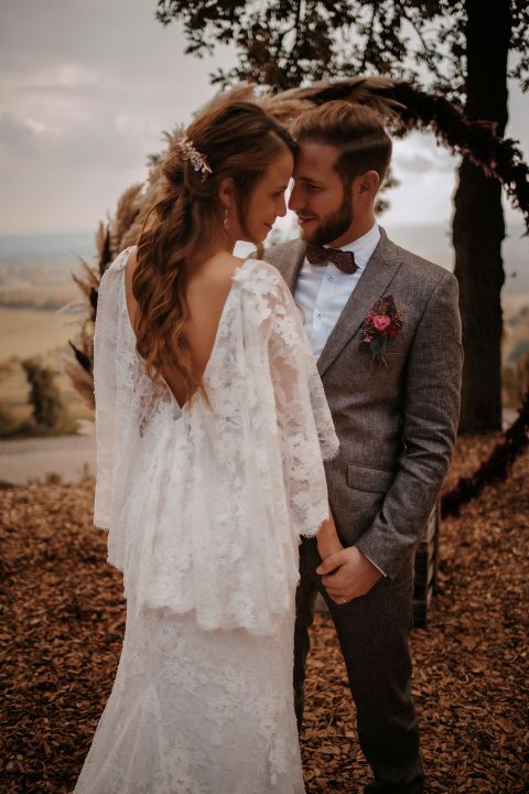 Hochzeitswahn - Braut und Bräutigam berühren sich intim die Stirn in einer ruhigen Umgebung im Freien, mit sanften Hügeln im Hintergrund. Sie trägt ein zartes Spitzenkleid und ein florales Haarteil, das Hochzeitseleganz verkörpert; er