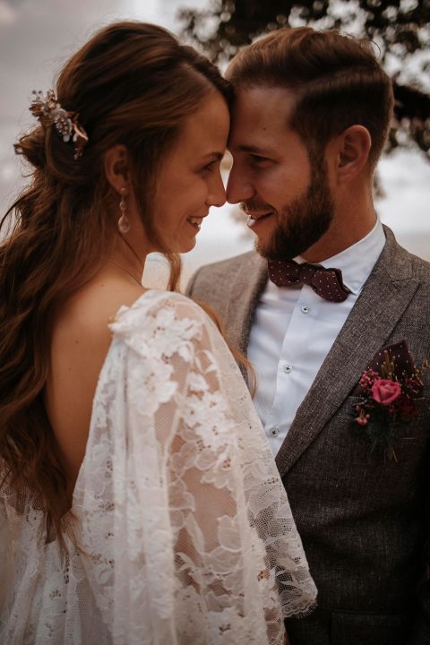 Hochzeitswahn - Braut und Bräutigam teilen einen zärtlichen Moment und lächeln sich eng an. Die Braut trägt einen Spitzenschleier und der Bräutigam einen grauen Anzug mit einer burgunderfarbenen Fliege und einem Blumenbouton