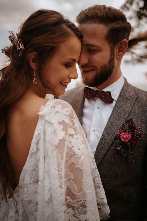 Hochzeitswahn - Braut und Bräutigam teilen einen zärtlichen Moment, lächeln sich innig an, während sie in Hochzeitskleidung gekleidet sind; die Braut in einem Spitzenkleid und der Bräutigam in einem Anzug mit Fliege und Blumenanstecker,