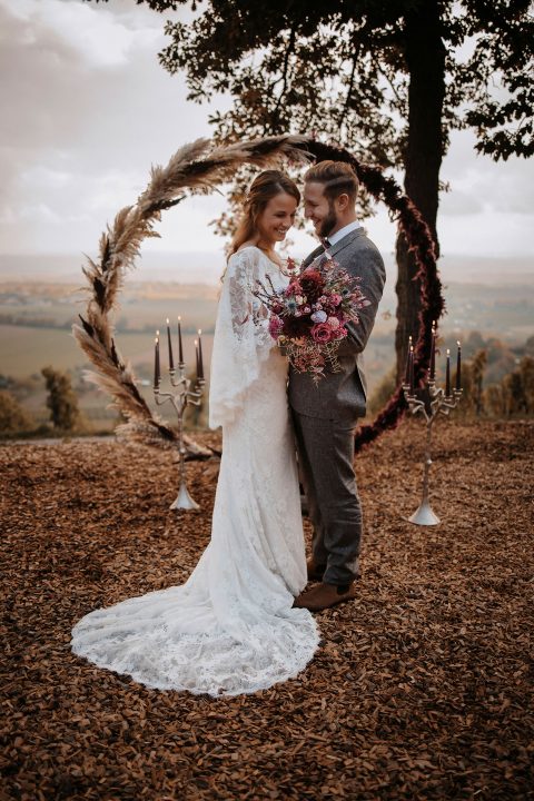Hochzeitswahn - Braut und Bräutigam verbringen einen intimen Moment unter einem rustikalen Blumenbogen in einer herbstlichen Umgebung im Freien, umgeben von gefallenen Blättern und einem malerischen Blick auf die Heuchelberger Warte. Die Braut hält ein lebendiges