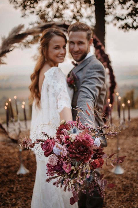 Hochzeitswahn - Ein frisch verheiratetes Paar posiert im Freien bei Sonnenuntergang. Die Braut, die einen farbenfrohen Blumenstrauß hält, lächelt neben dem Bräutigam im grauen Anzug. Beide sind von einer romantischen Kulisse mit Kerzen und einem Blumenbogen umgeben.