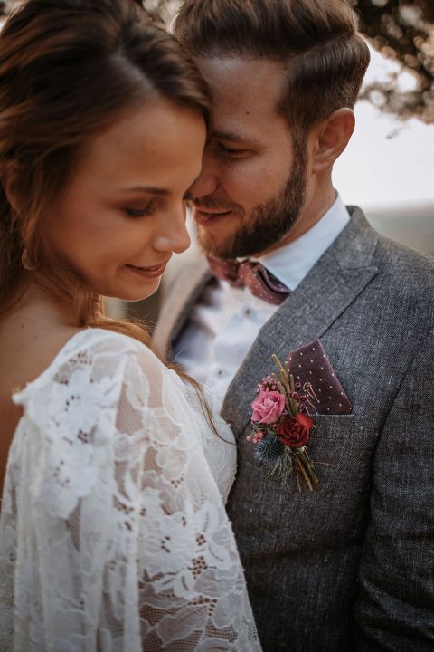 Hochzeitswahn - Ein Nahaufnahmebild von Braut und Bräutigam, die einen intimen Moment teilen. Die Braut, in einem elegant verzierten Spitzenkleid, lächelt sanft nach unten, während der Bräutigam, in einem grauen Anzug mit einem Blumenmuster