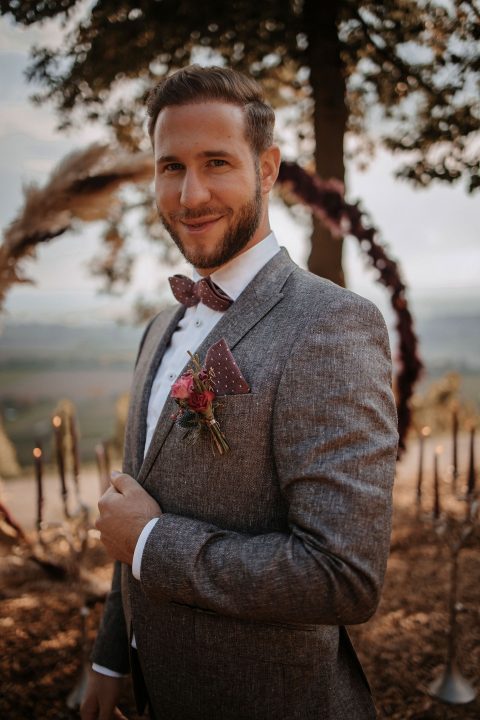 Hochzeitswahn - Ein Bräutigam in einem eleganten grauen Anzug mit Fliege lächelt in die Kamera und steht vor einem rustikalen Hochzeitsbogen mit einer malerischen Naturkulisse und verkörpert pure Hochzeitseleganz