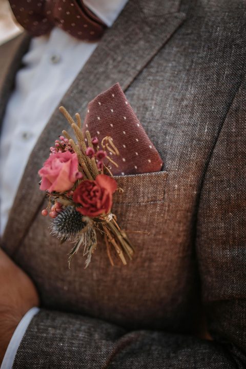Hochzeitswahn - Nahaufnahme eines braunen Tweed-Anzugssakkos mit weißem Hemd und burgunderfarbener gepunkteter Krawatte. Ein Einstecktuch und eine Blumenboutonniere mit rosa Blumen und kleinen Beeren verleihen Hochzeit