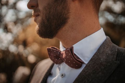 Hochzeitswahn - Nahaufnahme des unteren Gesichts und Halses eines Mannes, mit Bart und gepunkteter Fliege, im Hintergrund ein Fischgrätenanzug in weichem Fokus, der herbstliche