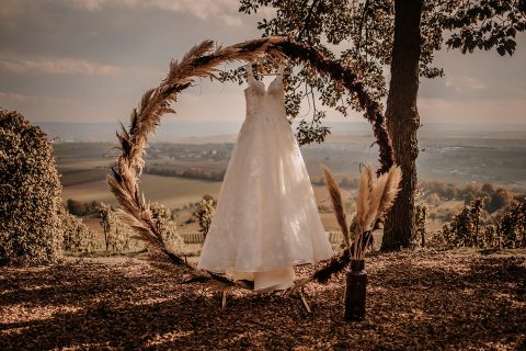 Hochzeitswahn - Ein mit herbstlicher Verspieltheit verziertes Brautkleid hängt an einem runden Blumenbogen auf einem Hügel mit Blick auf weite Felder unter einem wolkigen Himmel. Daneben stehen Federschmuck und eine rustikale Laterne.