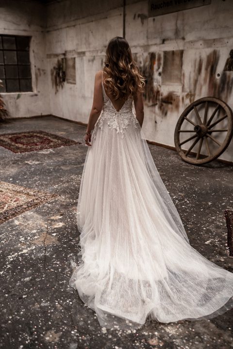 Hochzeitswahn - Eine Braut in einem eleganten Brautkleid mit Spitzenbesatz steht in einem rustikalen, verlassenen Raum mit dem Rücken zur Kamera, wodurch ein Kontrast zwischen dem zarten Kleid und der düsteren Umgebung entsteht.