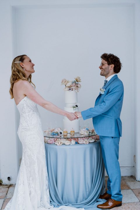 Hochzeitswahn - Ein fröhliches Paar im Hochzeitskleid hält Händchen und lacht gemeinsam neben einem Kuchentisch mit einer mehrstöckigen Torte und verschiedenen Cupcakes und feiert vor einem strahlend weißen Hintergrund einen Moment des Glücks.