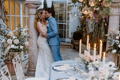 Hochzeitswahn - Ein Paar küsst sich zärtlich bei seiner Hochzeitsfeier im Kerzenschein, umgeben von eleganten Blumenarrangements und einem warm beleuchteten klassischen Veranstaltungsort, und fängt so einen Moment romantischer Feier ein.