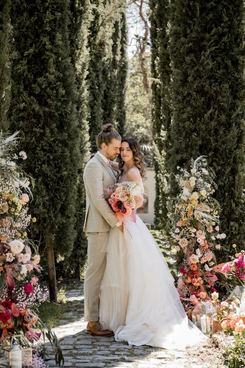 Hochzeitswahn - Ein Paar im Hochzeitskleid verbringt einen liebevollen Moment inmitten eines üppigen, mit Blumenarrangements geschmückten Gartengangs und genießt die Ruhe seines besonderen Tages.