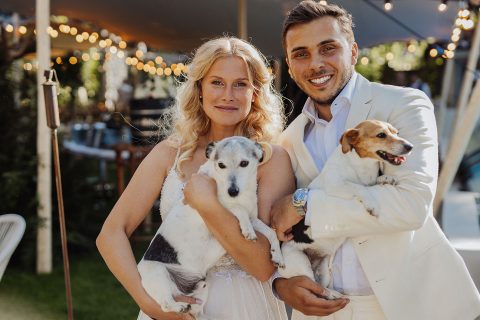 Hochzeitswahn - Eine Braut und ein Bräutigam lächeln bei einer Hochzeitsfeier im Freien und halten zwei kleine Hunde im Arm. Im Hintergrund hängen Lichterketten. Gut-Hohenholz