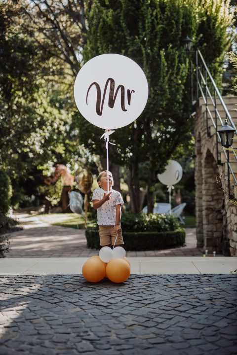 Hochzeitswahn - Ein Kind steht auf einem Kopfsteinpflasterweg und hält einen großen Luftballon mit der Aufschrift „Mr.“ in der Hand, flankiert von zwei kleineren orangefarbenen Luftballons, in einer sonnenbeschienenen Gartenlandschaft. Gut-Hohenholz