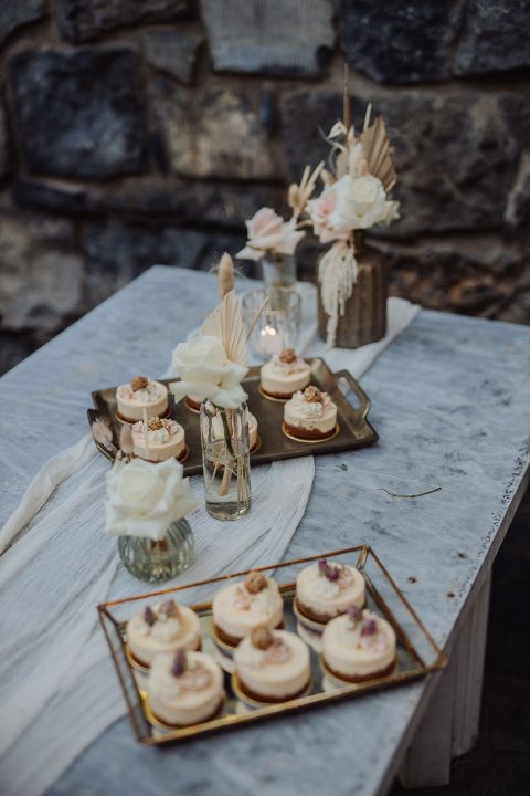 Hochzeitswahn - Ein rustikaler Desserttisch mit verschiedenen Gebäckstücken und einer Kerze als Tafelaufsatz vor einer Steinwand. Das Dekor umfasst Weiß- und Beigetöne mit subtilen floralen Akzenten. Gut-Hohenholz