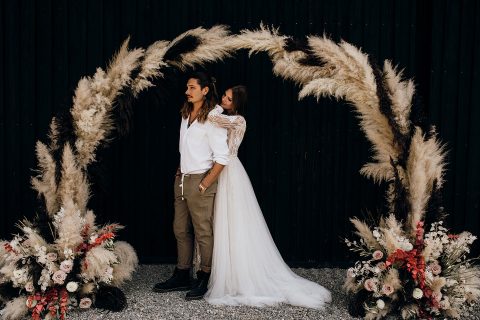 Hochzeitswahn - Ein Paar steht Hand in Hand unter einem mit Pampasgras und Blumen geschmückten Bogen und strahlt vor einem streng schwarzen Hintergrund eine unkonventionelle Eleganz aus.