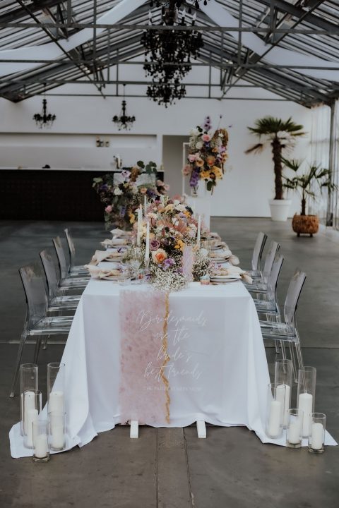 Hochzeitswahn - Elegante Tischdekoration für eine Hochzeit im Innenbereich mit einem langen weißen Tisch mit einem rosafarbenen Tischläufer in Ombre-Optik, garniert mit Blumengestecken, Kerzen und rundherum angeordneten durchsichtigen Stühlen in einem modernen Veranstaltungsort mit schwarzer Decke.