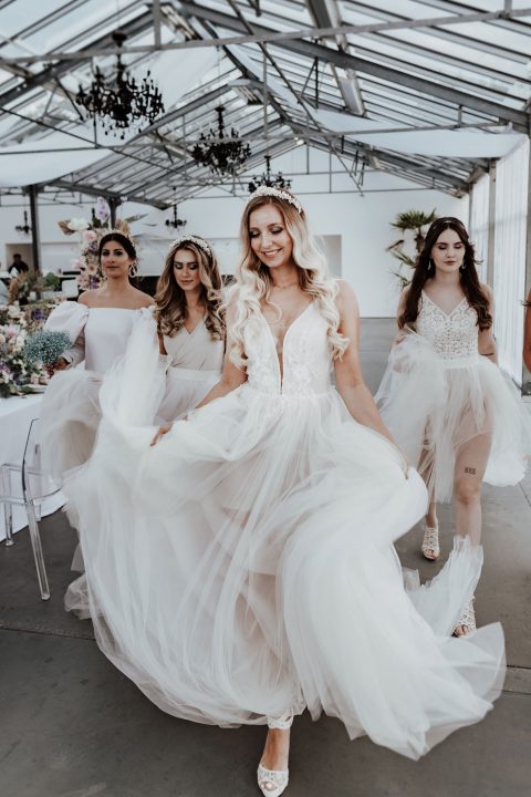 Hochzeitswahn - Eine Braut in einem fließenden weißen Kleid führt drei Brautjungfern in lavendelfarbenen Kleidern durch einen Veranstaltungsort im Industrie-Chic-Stil mit Kronleuchtern und eleganten Blumenarrangements.