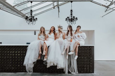 Hochzeitswahn - Fünf Frauen in weißen Kleidern und unterschiedlichen Frisuren sitzen nebeneinander auf einer Bank unter eleganten Kronleuchtern und verströmen eine schicke und festliche Atmosphäre, möglicherweise bei einer Hochzeitsfeier.