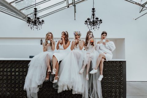 Hochzeitswahn - Vier Frauen in eleganten weißen Brautkleidern sitzen auf einer hohen Theke, jede hält eine Champagnerflasche und trägt bunte Blumenkränze. Sie befinden sich in einem modernen Raum, über dem stilvolle schwarze Kronleuchter hängen.