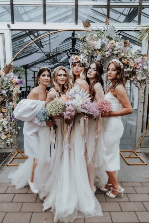 Hochzeitswahn - Fünf Frauen in eleganter Abendgarderobe halten große, bunte Blumensträuße in den Händen. Sie stehen zusammen in einem mit Blumen dekorierten Raum mit einer modernen Struktur aus Glas und Metall im Hintergrund.
