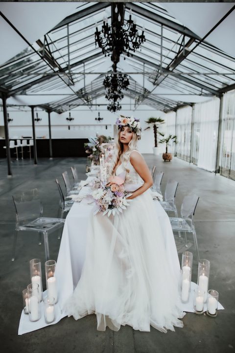 Hochzeitswahn - Eine Frau in einem weißen Brautkleid und mit Blumenkrone sitzt an einem langen Tisch in einem Raum mit Glaswänden und einem schwarzen Kronleuchter. Der Tisch ist mit einem weißen Tuch, üppigen Blumenarrangements und Kerzen in Glashaltern geschmückt.